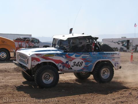 1990 Ford Bronco - white early Ford Bronco off-road race truck numbered 348 with stars-and-stripes graphics drives across a dirt course.