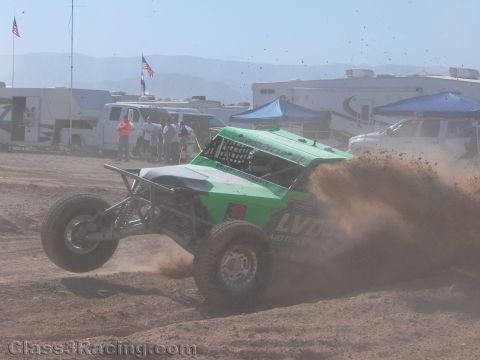 1990 Ford Bronco - bright green tube-chassis off-road race car kicks up a spray of dirt as it slides through a desert course lined with RVs and American flags.