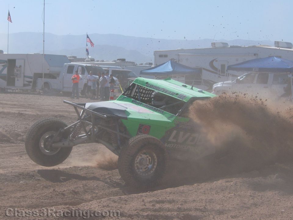 Green off-road race buggy throwing dirt rooster tail on desert course near RV pits