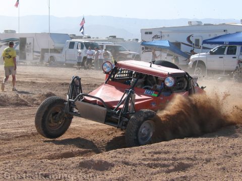 1990 Ford Bronco - red open-frame off-road race buggy number 1233 slides through loose dirt while spectators and motorhomes line the desert course behind it.