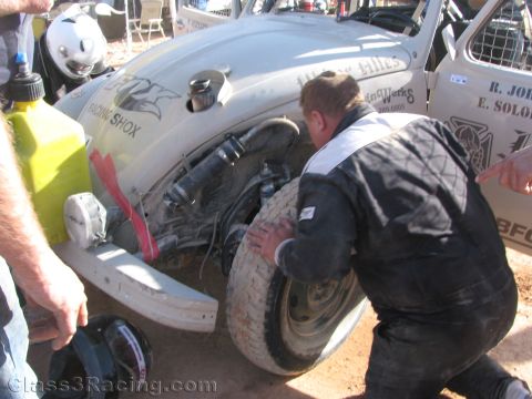 1990 Ford Bronco - crew member in dark coveralls crouches beside a white VW Beetle race car to work on a wheel, with a gas can and helmet nearby in the pits.