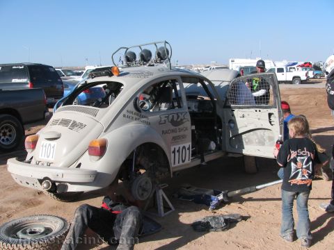 1990 Ford Bronco - white VW Beetle race car number 1101 sits on jack stands with its door open as crew work underneath and a child watches in the dirt pits.