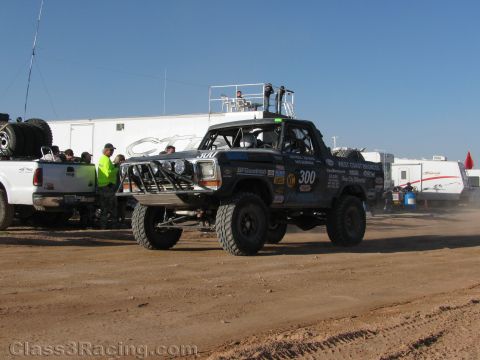1990 Ford Bronco - black Ford Bronco-style off-road race truck number 300 with light bar and sponsor decals is staged on dirt near trailers and RVs.