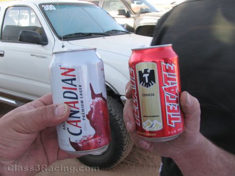 1990 Ford Bronco - Two hands hold up a Molson Canadian and a Tecate beer can in front of a white pickup truck at a dusty event.