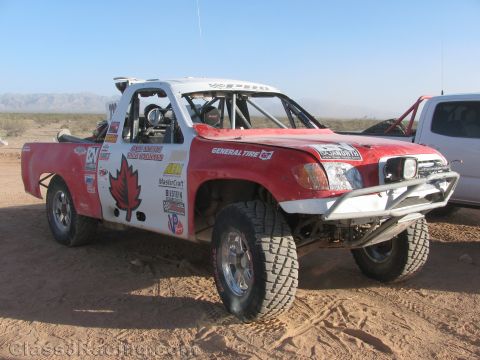 1990 Ford Bronco - red and white off-road race truck featuring a Canadian maple leaf graphic sits on desert sand with mountains in the distance.