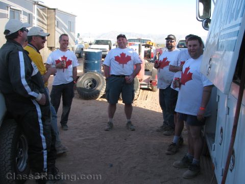1990 Ford Bronco - group of smiling men wearing matching white T-shirts with a red maple leaf stand and chat between trailers in a dusty desert camp.