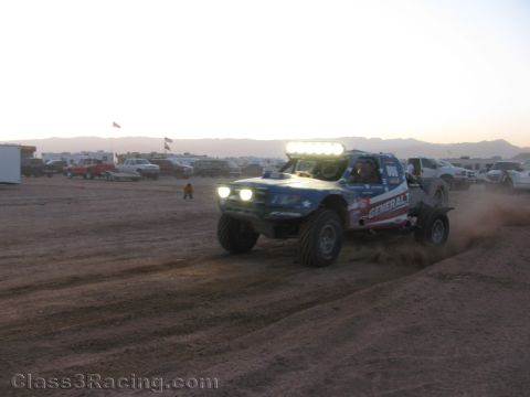 1990 Ford Bronco - off-road race truck with a glowing LED light bar and headlights kicks up dirt as it races across a desert course at dusk.