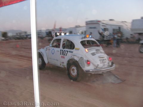 1990 Ford Bronco - white VW Beetle race car number 1107 drives through a dusty staging area at dusk, viewed past a tent pole and RVs.