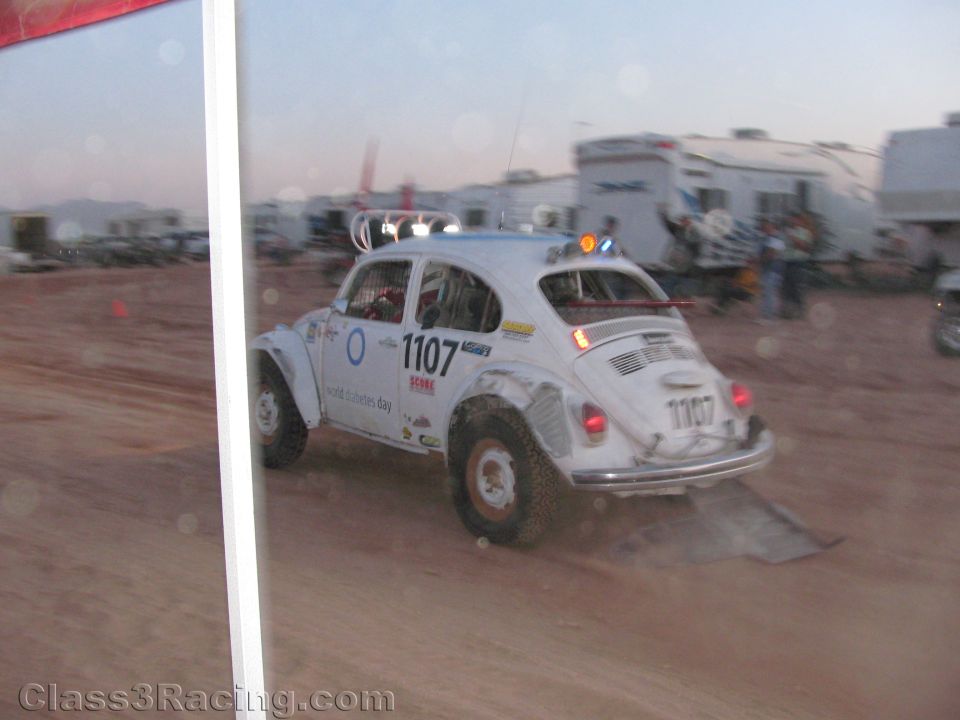 White Volkswagen Beetle race car number 1107 driving through dusty pits at dusk