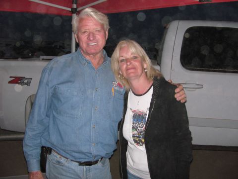 1990 Ford Bronco - smiling older man in a denim shirt poses with his arm around a woman in a graphic tee beneath a canopy, with a white pickup behind them.