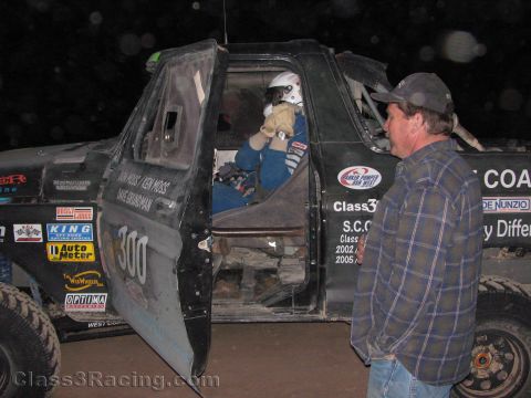 1990 Ford Bronco - man in a plaid shirt watches a helmeted driver climb into a battered black off-road race truck number 300 under night lighting.