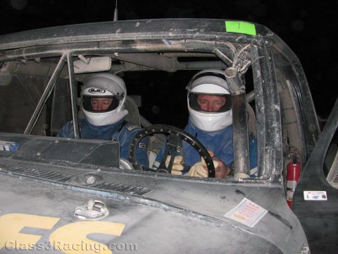 1990 Ford Bronco - Two drivers wearing helmets and blue racing suits sit side by side in the dusty caged cab of an off-road race truck at night.