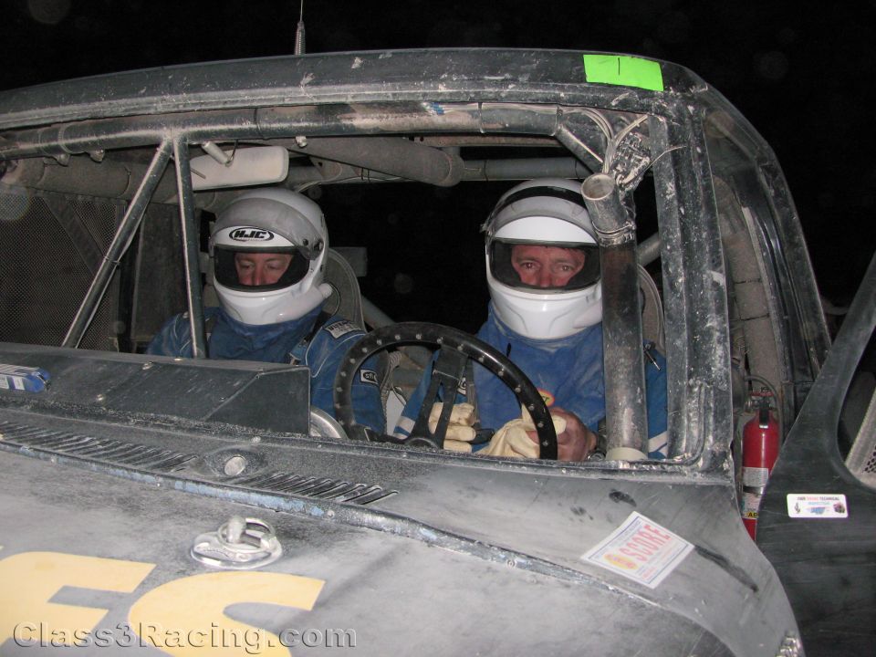 Two helmeted drivers seated side by side inside a dusty race truck cab at night