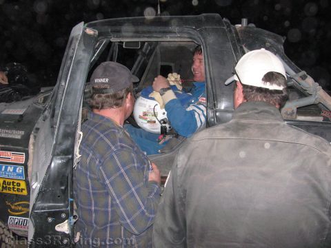 1990 Ford Bronco - Two crew members in caps lean into the cab of a black off-road race truck to talk with the helmeted driver under night lighting.