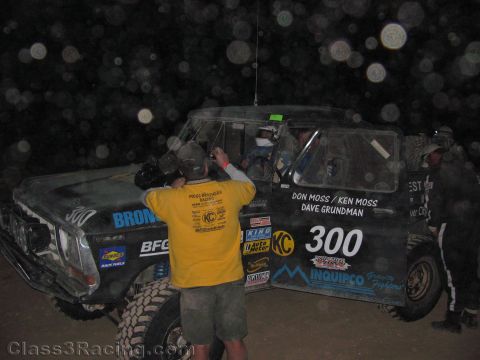 1990 Ford Bronco - crew member in a yellow shirt works on the dusty black Bronco-style race truck number 300 at night, sponsor decals visible on the door.