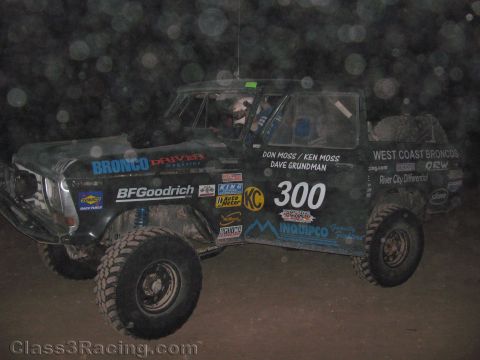 1990 Ford Bronco - dusty black Ford Bronco-style off-road race truck number 300 with West Coast Broncos decals sits side-on under night lighting.