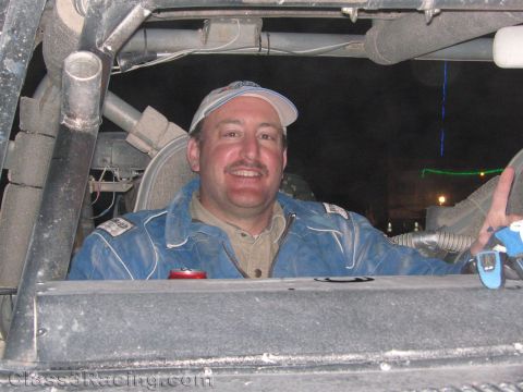 1990 Ford Bronco - smiling man wearing a cap and a worn blue racing suit sits inside the roll-caged interior of a race vehicle at night.