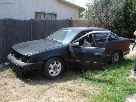 1989 Ford Taurus - A black sedan, likely a Ford Taurus from the mid-1980s, sits in a yard with its hood and grille missing.