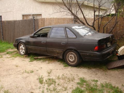 1989 Ford Taurus - A dark-colored sedan sits in a yard, partially supported by blocks, with a trailer ramp visible at the rear.