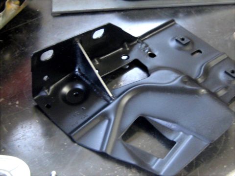 1996 Ford Bronco - A black-painted stamped-steel bracket sitting on a scratched shop bench, with an added gusset welded diagonally across its inner corner and…
