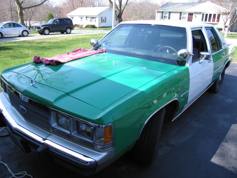 1991 Ford LTD Crown Victoria - green-and-white Ford sedan, with a pink cloth draped on its hood, parked in a residential driveway with houses behind.