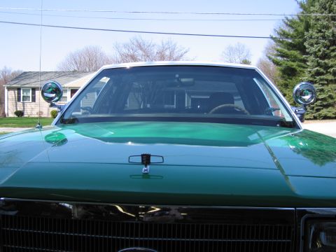1991 Ford LTD Crown Victoria - Close view over the hood and windshield of a green sedan fitted with two chrome side-mounted spotlights, house in background.