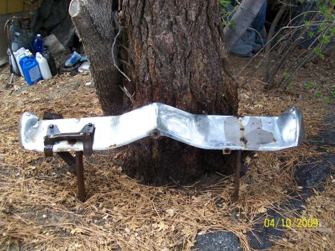 1973 Mercury Cougar - A chrome bumper, likely a replacement part, sits outdoors near a tree, showing signs of wear and tear.