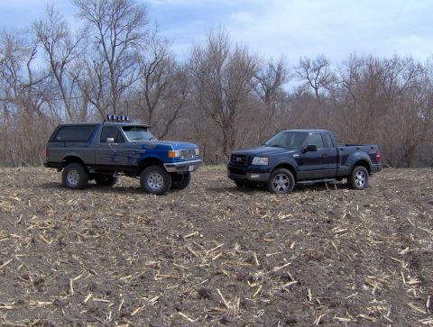 2005 Ford F-150 - A dark blue Ford F-150 pickup truck is parked next to a lifted blue Bronco in a field with bare trees.