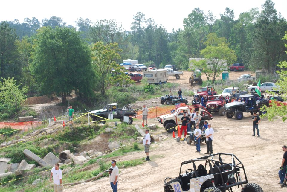 Group of off-road vehicles and people gathered at an outdoor event, with trees and a dirt track visible