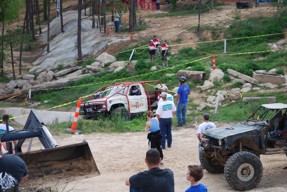 Two off-road trucks are parked on a dirt track with spectators around. One truck is white with a red roll cage, the other is dark colored