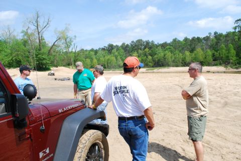 2008 Jeep Wrangler - Group of men gathered around a red Jeep Wrangler on a sandy beach with trees in the background.