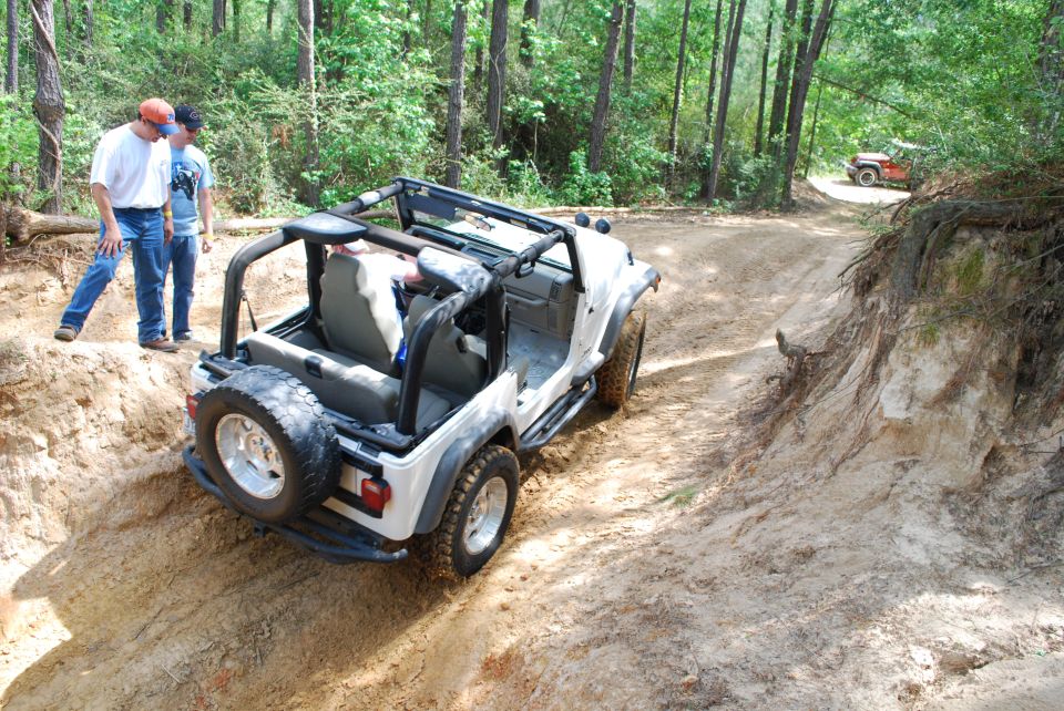 White Jeep Wrangler on a dirt trail with two people standing nearby; another red Jeep is visible in the distance