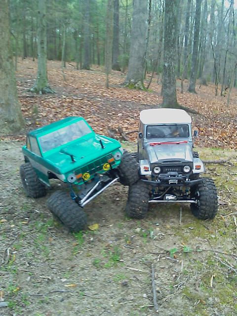 1996 Ford Bronco - Two RC trucks, one green and one silver, are positioned in a forest clearing.