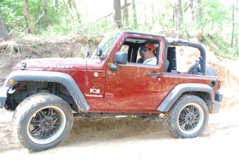 2008 Jeep Wrangler - A red Jeep Wrangler is shown from the side, driving off-road on a dirt trail with trees in the background.