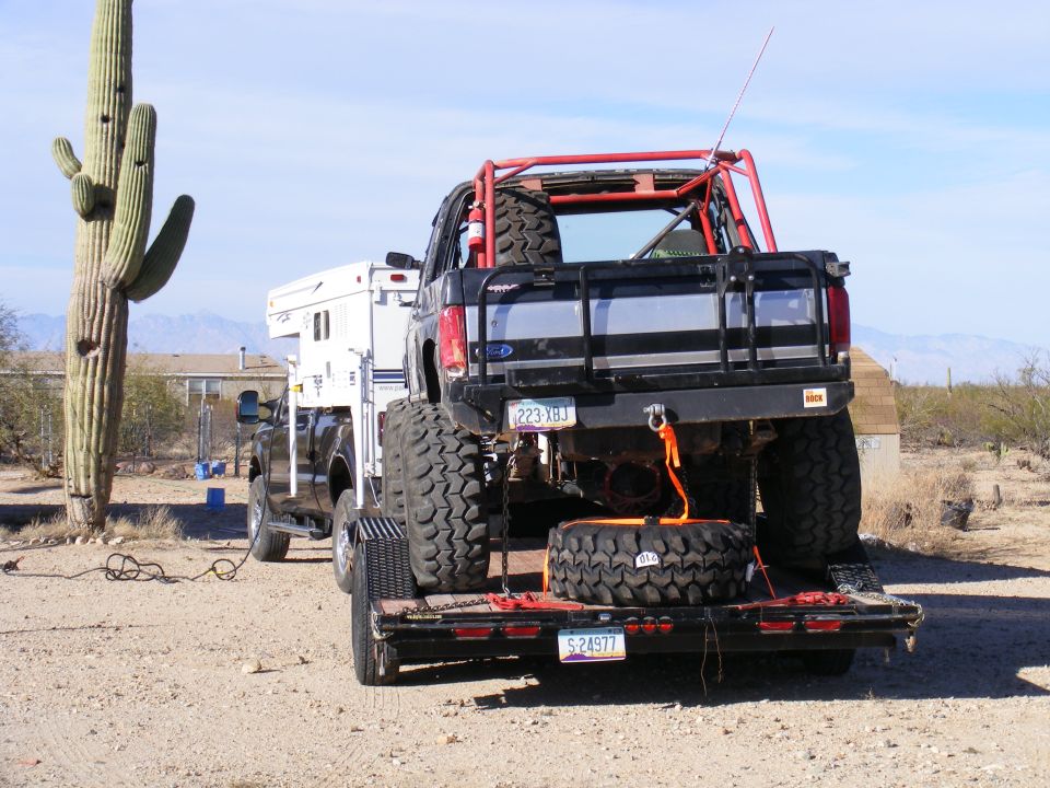 Black lifted Ford pickup with red roll cage and big mud tires on a trailer, towed by a truck with camper in the desert