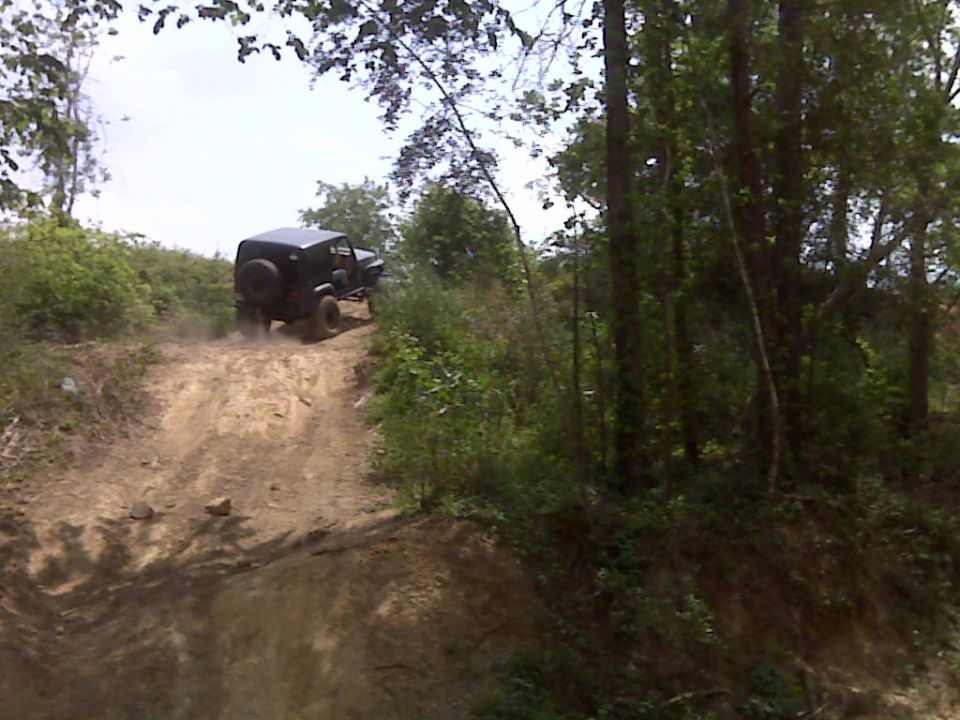 Black Jeep Wrangler driving up a steep dirt hill, kicking up dust. Trees and foliage surround the path