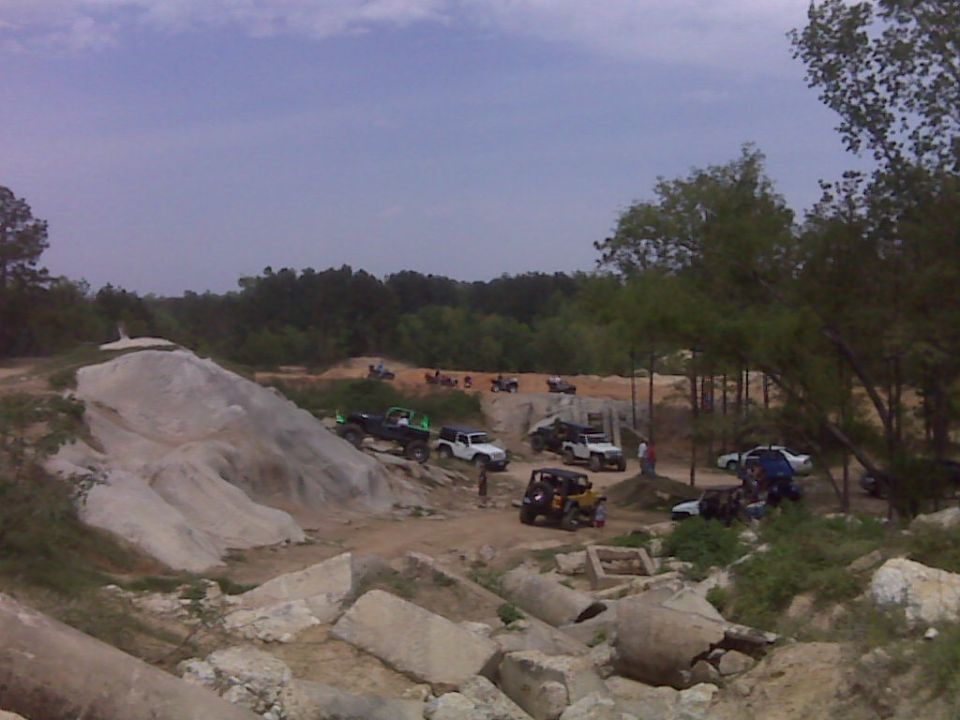 Several off-road vehicles, including Jeeps and trucks, gathered at a dirt park with rocky terrain and trees