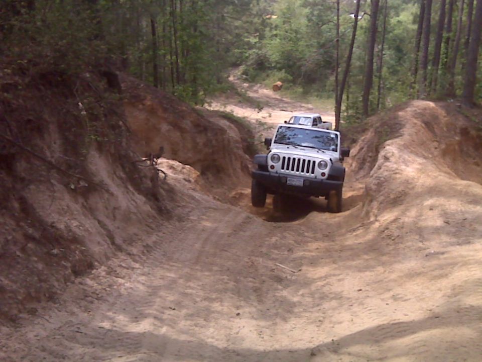 White Jeep Wrangler driving on a steep dirt trail with trees in the background. Another vehicle is visible behind it