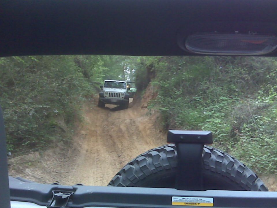 Rear window view past spare tire of a white Jeep Wrangler JK following on a dirt trail