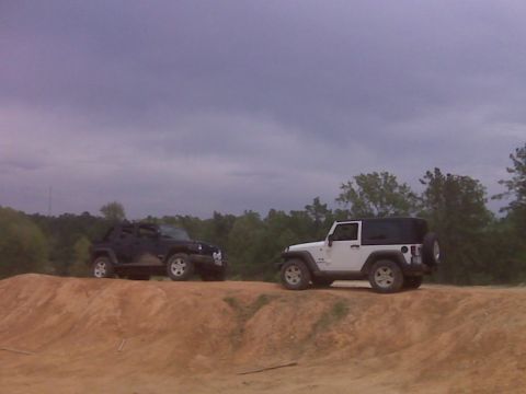 2008 Jeep Wrangler - Two Jeep Wranglers are positioned on a dirt embankment, with trees in the background and a cloudy sky above.