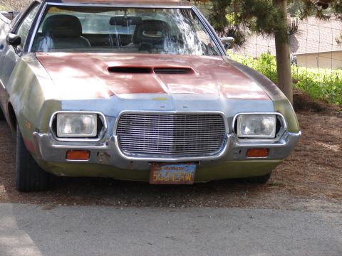 1973 Ford Ranchero - Head-on front view of a rough project Ford Ranchero-style car car in patchy primer, faded green paint and bare metal, wearing a hood with a…