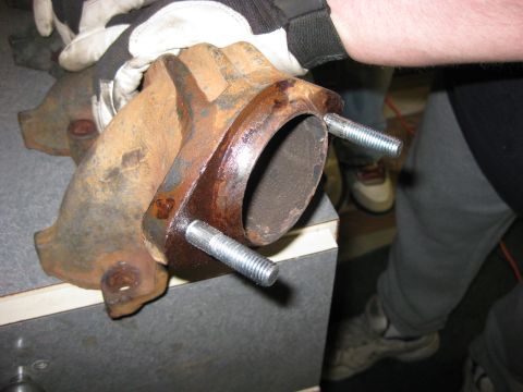 1979 Ford Bronco - A gloved hand holds a rusty cast-iron exhaust manifold up to the camera, focused on the collector flange with two new zinc-plated studs thr…