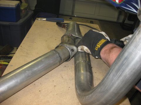 1979 Ford Bronco - Bare steel tubing being test-fitted on a plywood workbench in a garage, with a gloved hand (black 'TrueFit' glove) holding a notched tube a…