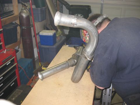 1979 Ford Bronco - A fabricator in a welding hood and blue work shirt tack-welds a bare-steel tube assembly clamped on a plywood workbench in a home garage, w…