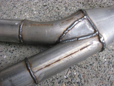 1979 Ford Bronco - Close-up of a fabricated bare-steel tube assembly lying on concrete, showing a Y-shaped junction where two bent tubes are joined with a gus…