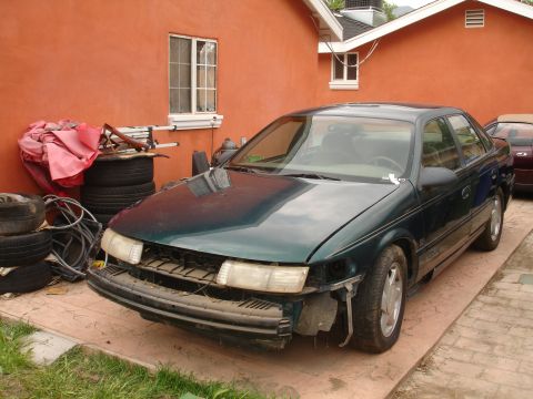1995 Ford Taurus - A green sedan sits in a driveway with its front bumper missing, revealing the internal components.