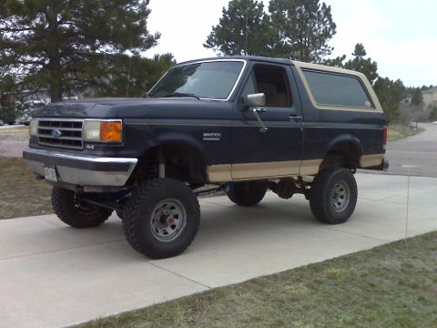 1990 Ford Bronco - This lifted 1990 Ford Bronco sits on a paved driveway with trees in the background.