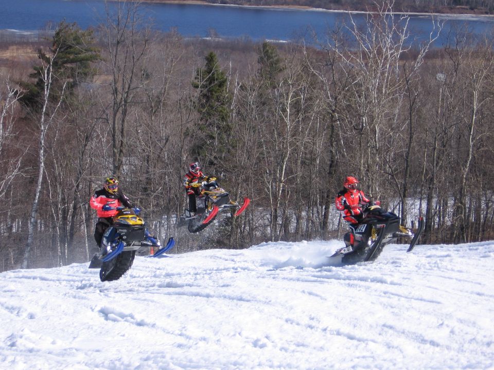 Three snowmobiles jumping a snow hill side by side with a lake in the background