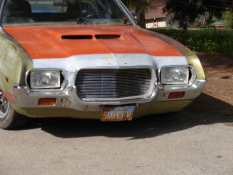1973 Ford Ranchero - Close front-end view of a gold/olive 1970s Ford intermediate with a primered orange-and-white striped hood, twin hood scoops, quad stacked…