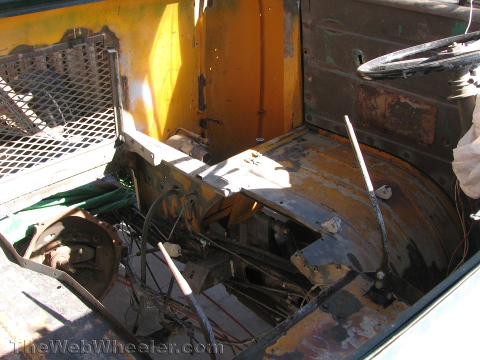 Disassembled vehicle interior showing rusted floor pan, steering wheel, and exposed metalwork
